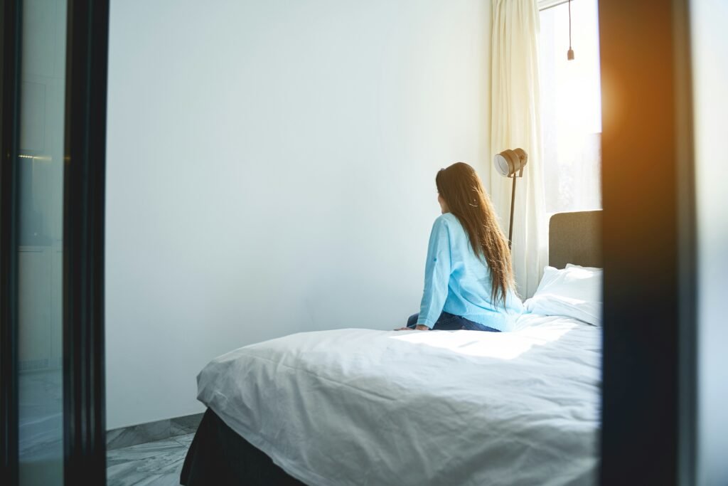 Woman sitting alone on the edge of a bed, looking toward a sunlit window