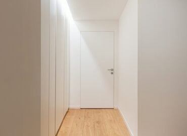 Closed door at the end of a quiet sunlit hallway, representing a young adult withdrawn during Failure to Launch