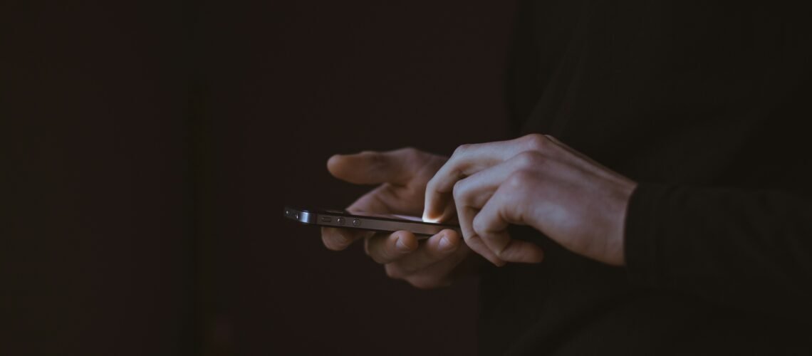 Hands holding a phone in darkness, the screen as the only source of light — illustrating compulsive phone use and screen dependency.