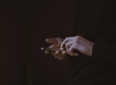 Hands holding a phone in darkness, the screen as the only source of light — illustrating compulsive phone use and screen dependency.