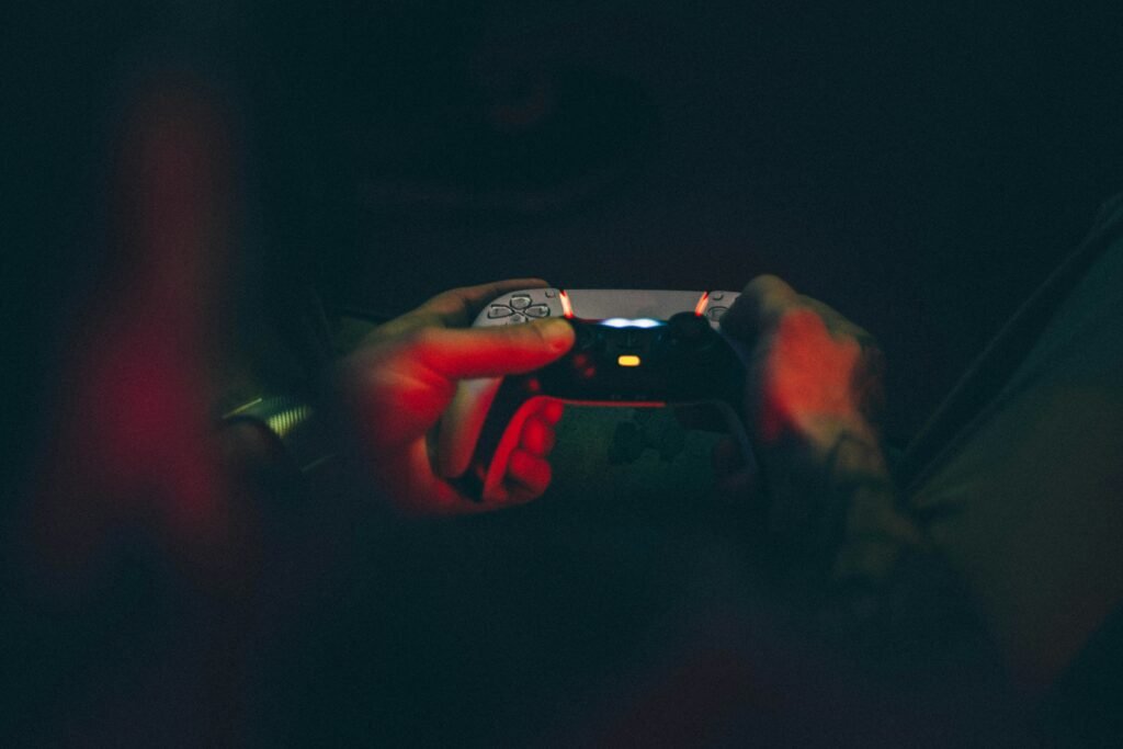 Hands gripping a game controller in near-total darkness, the screen and controller lights illuminating the late-night isolation of gaming addiction.